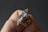 A hand holds an intricately designed ring featuring an iridescent stone set in a detailed metal framework. The metal has an elaborate, wire-like design that spirals around the stone.