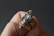 A hand holds an intricately designed ring featuring an iridescent stone set in a detailed metal framework. The metal has an elaborate, wire-like design that spirals around the stone.