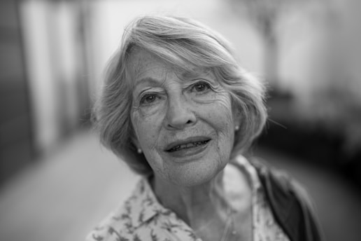 An elderly woman with short, wavy hair looks directly at the camera with a gentle expression. Her features are highlighted by the soft lighting, and she is wearing a patterned blouse. The background is blurred, drawing attention to her face.