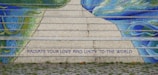 Mosaic steps featuring vibrant shades of blue and green create an artistic pattern. A motivational message in bold capitals reads 'RADIATE YOUR LOVE AND UNITY TO THE WORLD.' The foreground includes a cobblestone path and patches of grass.