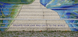 Mosaic steps featuring vibrant shades of blue and green create an artistic pattern. A motivational message in bold capitals reads 'RADIATE YOUR LOVE AND UNITY TO THE WORLD.' The foreground includes a cobblestone path and patches of grass.