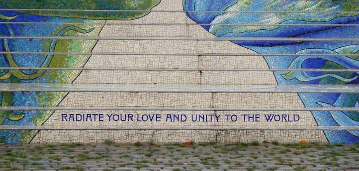 Mosaic steps featuring vibrant shades of blue and green create an artistic pattern. A motivational message in bold capitals reads 'RADIATE YOUR LOVE AND UNITY TO THE WORLD.' The foreground includes a cobblestone path and patches of grass.