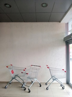 Three empty shopping carts with red handles are positioned against a light beige wall. The carts are placed on a tiled floor under a dark ceiling with spotlights. A glass door on the right lets in natural light.