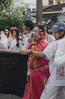 A vibrant street scene features participants in traditional attire, including a person in bright pink clothing with ceremonial decorations on their face. Several people walk closely, carrying banners with intricate designs. A festive atmosphere is present, with greenery and urban elements in the background.