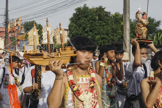A group of individuals participate in a cultural parade or festival. The image captures several young men, one of whom is prominently holding a large wooden ship model. Another individual is carrying an ornate figure with traditional attire. They wear intricate costumes adorned with garlands of flowers, and some have facial piercings. The background shows trees and possibly a temple structure, indicating an outdoor setting.