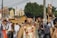 A group of individuals participate in a cultural parade or festival. The image captures several young men, one of whom is prominently holding a large wooden ship model. Another individual is carrying an ornate figure with traditional attire. They wear intricate costumes adorned with garlands of flowers, and some have facial piercings. The background shows trees and possibly a temple structure, indicating an outdoor setting.