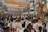 A vibrant street procession with numerous people wearing white attire, many of whom are carrying religious artifacts. Colorful banners hang above the street, written in what appears to be a Southeast Asian script. The atmosphere is lively, with several stalls lining the street and a visible sense of community and celebration.