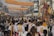 A vibrant street procession with numerous people wearing white attire, many of whom are carrying religious artifacts. Colorful banners hang above the street, written in what appears to be a Southeast Asian script. The atmosphere is lively, with several stalls lining the street and a visible sense of community and celebration.