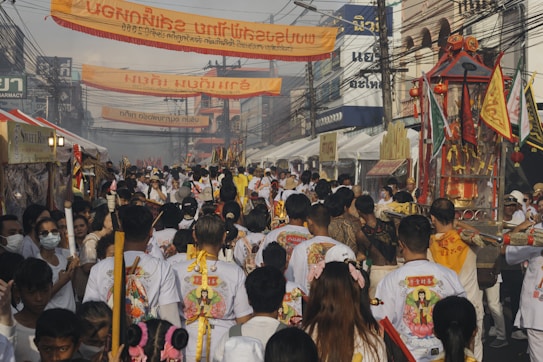 A vibrant street procession with numerous people wearing white attire, many of whom are carrying religious artifacts. Colorful banners hang above the street, written in what appears to be a Southeast Asian script. The atmosphere is lively, with several stalls lining the street and a visible sense of community and celebration.