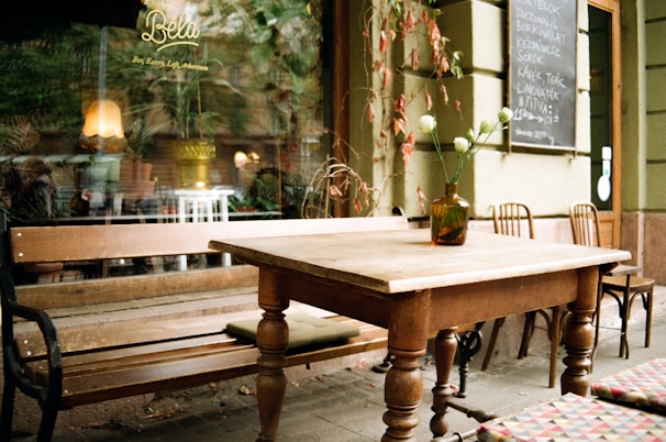 Cozy outdoor café setting with a wooden table and bench, decorated with a vase of white flowers. The background offers a glimpse of the interior through a window, showing a warm-lit lamp and plants. A chalkboard menu is on the wall, adding to the quaint atmosphere.