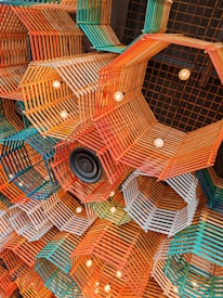 A complex and colorful ceiling installation composed of geometric wooden frames in orange, teal, and white hues. The arrangement includes exposed light bulbs embedded within the frames, along with a black speaker, adding an artistic and modern touch.