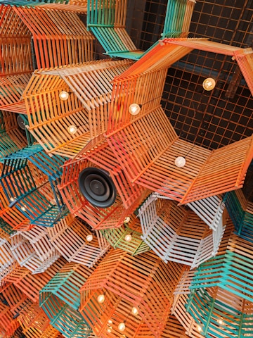A complex and colorful ceiling installation composed of geometric wooden frames in orange, teal, and white hues. The arrangement includes exposed light bulbs embedded within the frames, along with a black speaker, adding an artistic and modern touch.