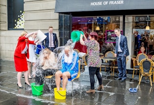 Several people are participating in the ice bucket challenge outside a building with 'House of Fraser' signage. Two seated individuals are being drenched with water from buckets by others standing around them. The scene includes wet pavement and café chairs nearby, and the expressions are lively, suggesting enjoyment and participation in a social activity.
