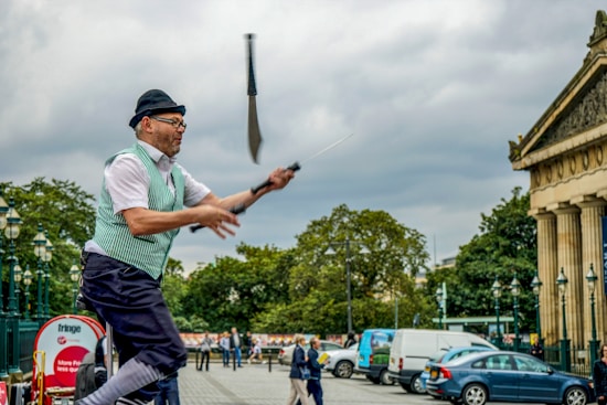 A performer in a green vest and black hat is juggling knives in an outdoor urban setting. The scene includes a mix of historical and modern elements, with a classical building on the right and cars parked along the street. Several people are walking in the background under a cloudy sky.