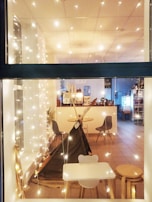 A cozy indoor setting viewed through a window, decorated with string lights that create a warm and inviting atmosphere. Inside, there is a small play tent resembling a teepee, surrounded by modern chairs and a small table with stools. The floor is tiled and the room has a simple, clean design with a white color scheme. In the background, there are additional chairs and tables, as well as a counter with a water dispenser and some plants.