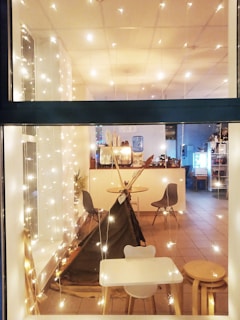 A cozy indoor setting viewed through a window, decorated with string lights that create a warm and inviting atmosphere. Inside, there is a small play tent resembling a teepee, surrounded by modern chairs and a small table with stools. The floor is tiled and the room has a simple, clean design with a white color scheme. In the background, there are additional chairs and tables, as well as a counter with a water dispenser and some plants.