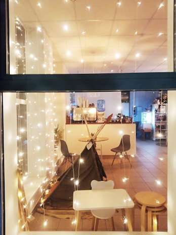 A cozy indoor setting viewed through a window, decorated with string lights that create a warm and inviting atmosphere. Inside, there is a small play tent resembling a teepee, surrounded by modern chairs and a small table with stools. The floor is tiled and the room has a simple, clean design with a white color scheme. In the background, there are additional chairs and tables, as well as a counter with a water dispenser and some plants.