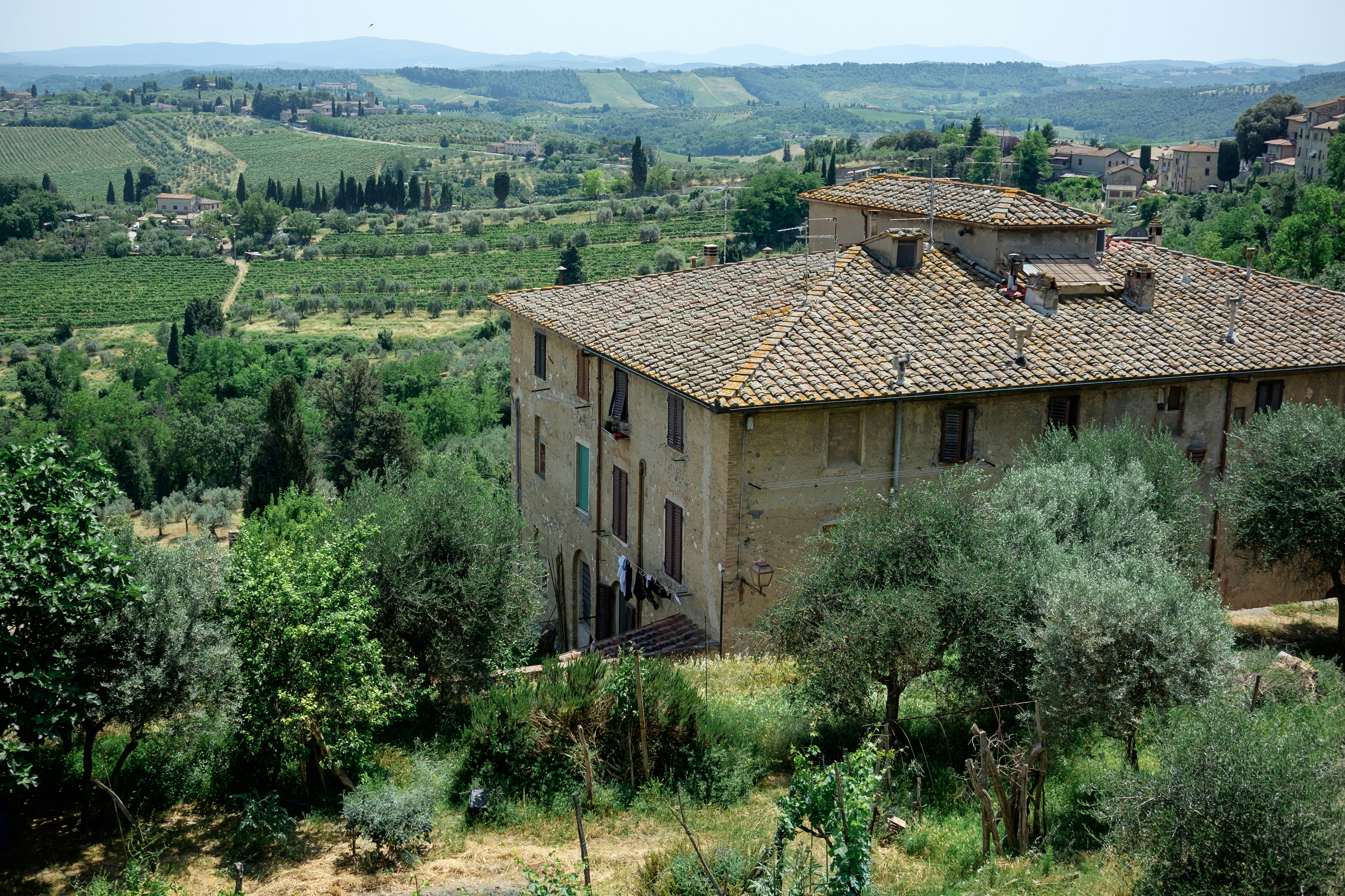 A rustic, stone building with a traditional tiled roof is set in a lush, green landscape. The surrounding area includes dense vegetation, trees, and rolling hills extending into the distance. The atmosphere is serene and idyllic, providing a panoramic view of the countryside with scattered farmhouses and a distant hill range.