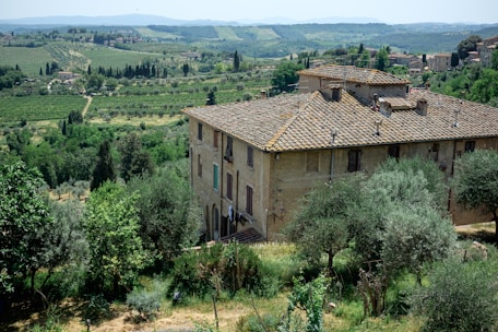 A rustic, stone building with a traditional tiled roof is set in a lush, green landscape. The surrounding area includes dense vegetation, trees, and rolling hills extending into the distance. The atmosphere is serene and idyllic, providing a panoramic view of the countryside with scattered farmhouses and a distant hill range.