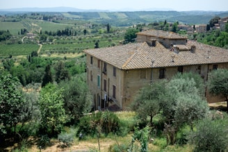 A rustic, stone building with a traditional tiled roof is set in a lush, green landscape. The surrounding area includes dense vegetation, trees, and rolling hills extending into the distance. The atmosphere is serene and idyllic, providing a panoramic view of the countryside with scattered farmhouses and a distant hill range.