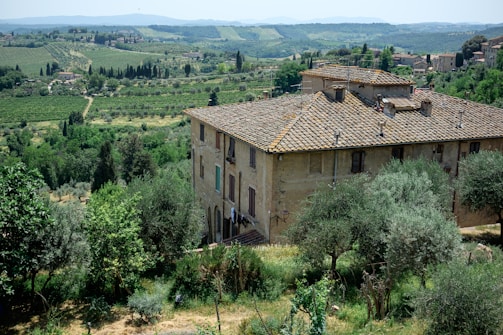 A rustic, stone building with a traditional tiled roof is set in a lush, green landscape. The surrounding area includes dense vegetation, trees, and rolling hills extending into the distance. The atmosphere is serene and idyllic, providing a panoramic view of the countryside with scattered farmhouses and a distant hill range.