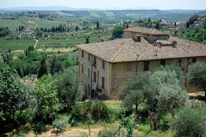 A rustic, stone building with a traditional tiled roof is set in a lush, green landscape. The surrounding area includes dense vegetation, trees, and rolling hills extending into the distance. The atmosphere is serene and idyllic, providing a panoramic view of the countryside with scattered farmhouses and a distant hill range.