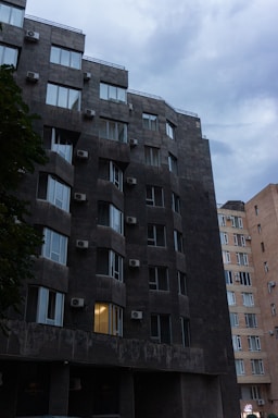 A dark, modern apartment building with multiple windows and air conditioning units is pictured. The sky is overcast, contributing to a gloomy atmosphere. One window is illuminated with a warm light, contrasting with the otherwise dark facade. Adjacent to this building, there is a lighter-colored building with a similar architectural style.