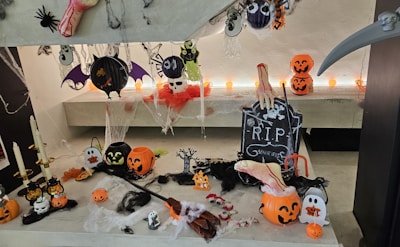 An array of Halloween decorations is arranged, featuring various elements like pumpkins with carved faces, a tombstone with 'R.I.P' inscribed, hanging bats, skulls, and ghosts. A bloody hand prop, spiders, and cotton webs add to the spooky atmosphere. Candles and orange string lights provide illumination, enhancing the eerie setting.