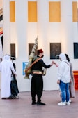 A street vendor wearing traditional clothing delivers a drink to a woman in casual attire. The vendor carries a large, ornate brass container on his back. Other individuals, possibly tourists or locals, are nearby, some dressed in traditional Middle Eastern garments. They stand in front of a building with large white and yellow columns.