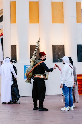 A street vendor wearing traditional clothing delivers a drink to a woman in casual attire. The vendor carries a large, ornate brass container on his back. Other individuals, possibly tourists or locals, are nearby, some dressed in traditional Middle Eastern garments. They stand in front of a building with large white and yellow columns.
