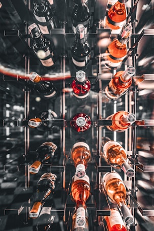 Rows of wine bottles are arranged horizontally on metal racks. The bottles have various colors of liquid, including red, rose, and white, with labels visible on some. The overall atmosphere is elegant and well-organized, suggesting a wine cellar or a specialized storage area.