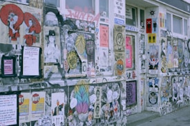 A wall covered with a diverse array of posters, graffiti, and street art. Various images, illustrations, and texts are layered over each other, creating a collage effect. Some posters feature stylized portraits and symbolic icons, while others incorporate text or slogans. The overall aesthetic is eclectic and vibrant.