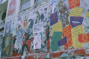 A wall filled with various posters and graffiti in multiple colors. Some posters include messages like 'Your right to privacy has been revoked,' 'There's no app for this,' and 'Take sharing offline.' Artistic elements include a cartoonish skeleton figure and various abstract faces. The scene shows a mix of street art and political or social commentary.