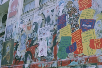 A wall filled with various posters and graffiti in multiple colors. Some posters include messages like 'Your right to privacy has been revoked,' 'There's no app for this,' and 'Take sharing offline.' Artistic elements include a cartoonish skeleton figure and various abstract faces. The scene shows a mix of street art and political or social commentary.