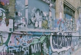 A brick wall is covered with vibrant graffiti, including tags, stickers, and abstract art. The artwork displays a mix of bold colors and intricate designs, with windows partially showing behind the layers of graffiti. The wall has a raw urban aesthetic.
