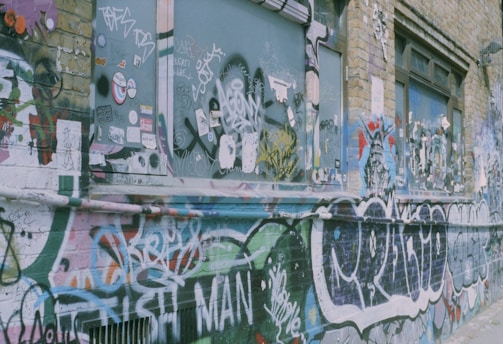 A brick wall is covered with vibrant graffiti, including tags, stickers, and abstract art. The artwork displays a mix of bold colors and intricate designs, with windows partially showing behind the layers of graffiti. The wall has a raw urban aesthetic.