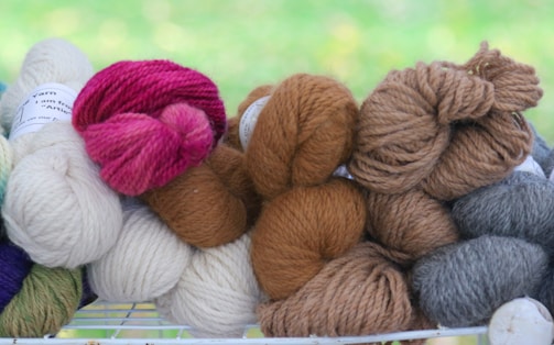 A collection of yarn skeins in different colors, including cream, pink, brown, and gray, arranged together on a wire rack. The background appears to be a blurred green space, possibly grass or foliage.