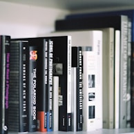 A collection of photography books neatly arranged on a shelf. The titles visible include 'Humans of New York,' 'The Polaroid Book,' 'Icons of Photography,' and 'The Photographer's Eye.' The books are of varying sizes and colors, predominantly in shades of black, white, and gray.