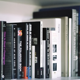 A collection of photography books neatly arranged on a shelf. The titles visible include 'Humans of New York,' 'The Polaroid Book,' 'Icons of Photography,' and 'The Photographer's Eye.' The books are of varying sizes and colors, predominantly in shades of black, white, and gray.