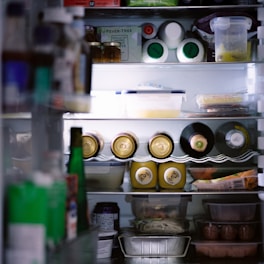 An organized refrigerator filled with various items, including bottles, cans, and containers. The top shelves hold jars and cartons, while the middle section contains several canned drinks and bottles of wine or sparkling water. The lower shelves have food containers, possibly containing leftovers or prepared meals.
