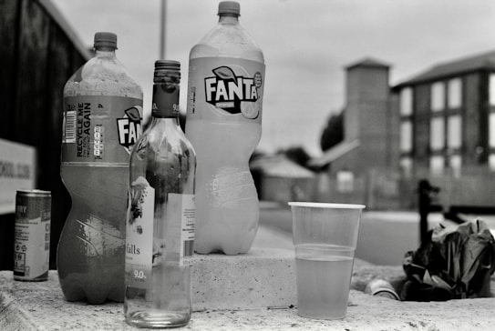 A collection of empty and partially filled beverage containers is placed on a concrete surface. Among them are two large Fanta bottles, a clear wine bottle, a small aluminum can, and a plastic cup filled with liquid. In the background, a blurred urban scene features a few buildings and a cloudy sky.