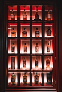 A wooden display case containing multiple bottles of whiskey, each illuminated by a warm light. The shelves are organized in a grid with three rows and four columns, showcasing various brands such as Balblair and Old Pulteney. The background lighting provides a cozy ambiance, highlighting the amber color of the whiskey.