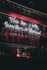A dimly lit bar with shelves filled with bottles. Neon signage hangs above the bottles, creating a retro and warm atmosphere. Glassware is placed on the counter below.