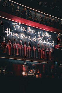 A dimly lit bar with shelves filled with bottles. Neon signage hangs above the bottles, creating a retro and warm atmosphere. Glassware is placed on the counter below.