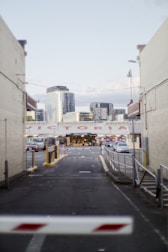 An urban alleyway leads to a market entrance with the word 'WELCOME' displayed prominently. Surrounding buildings include modern skyscrapers in the background and brick structures flanking the alley. Cars are parked along the alleyway, and a barrier gate is partially visible at the foreground.