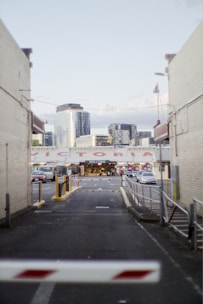 An urban alleyway leads to a market entrance with the word 'WELCOME' displayed prominently. Surrounding buildings include modern skyscrapers in the background and brick structures flanking the alley. Cars are parked along the alleyway, and a barrier gate is partially visible at the foreground.