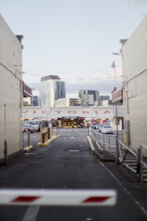 An urban alleyway leads to a market entrance with the word 'WELCOME' displayed prominently. Surrounding buildings include modern skyscrapers in the background and brick structures flanking the alley. Cars are parked along the alleyway, and a barrier gate is partially visible at the foreground.