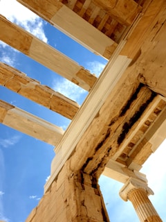 The image captures a view of ancient limestone ruins against a clear blue sky. The architectural structure is defined by large stone blocks with visible aging and wear. The perspective focuses on the details of the columns and beams, highlighting the intricate design and natural erosion over time.