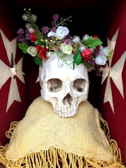 A skull rests on a cushioned surface adorned with an ornate garland of colorful flowers and green leaves. The background is rich red decorated with white star-like patterns.