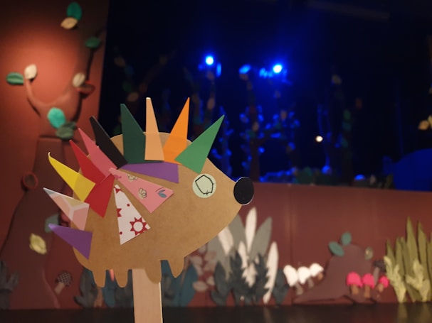 A creatively designed hedgehog puppet with colorful, geometric paper spikes is in the foreground. The background features a stage with a woodland-themed set design including leaves and plants, illuminated with blue stage lights.
